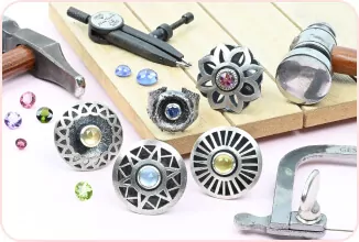 Metal jewelry with colored gems on a wooden board, surrounded by tools like a hammer and ring sizer.