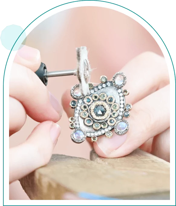 Hands polishing a detailed, ornate pendant with a tool, featuring gemstones and intricate metalwork.
