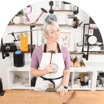 Person in a workshop holding a hammer, standing by a workbench, wearing an apron. Shelves with tools in background.