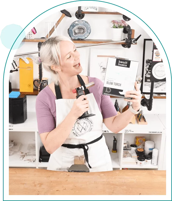 Person holding a jewelers torch and its packaging, smiling in a jewelry making workshop with various tools on shelves behind.