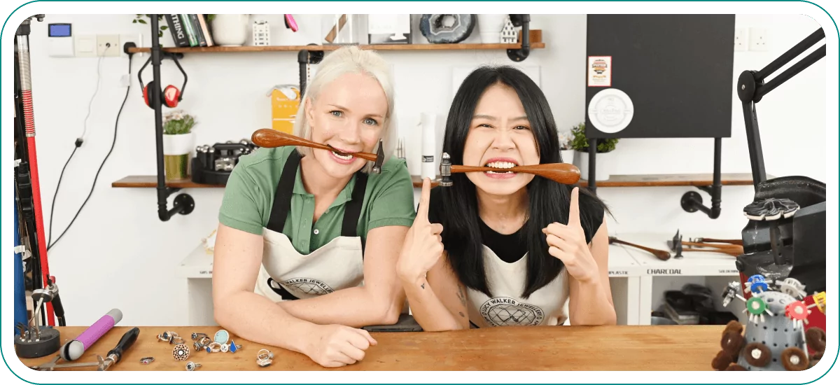 Two women smiling, holding jewelers hammers their mouths, sitting at a jewelers table with various supplies.