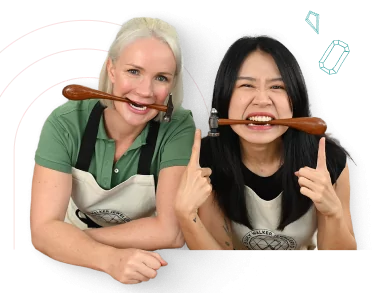 Two women smiling, holding jewelers hammers their mouths, pointing upwards, wearing aprons.  From a class on how to make silver hoop earrings.