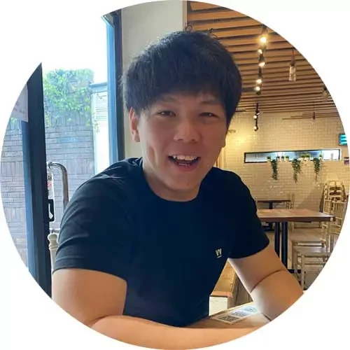 Man smiling at a table in a cafe with wooden ceiling and hanging lights in the background.