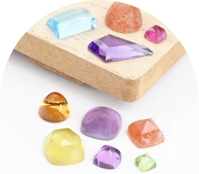 Various colorful gemstones for jewelry making on and near a wooden block, including blue, purple, pink, orange, yellow, and green stones.