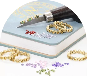 Gold rings on a book with scattered colorful gemstones and a graver for stone setting.
