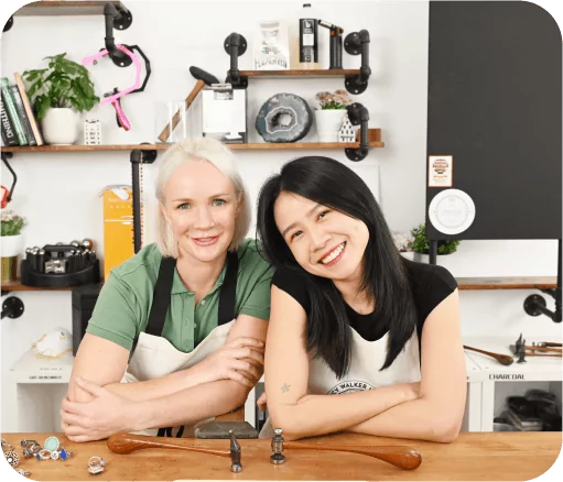 Two women smiling, wearing aprons, sitting at a jewelers workbench with jewelry making tools, shelves, and various items in the background.