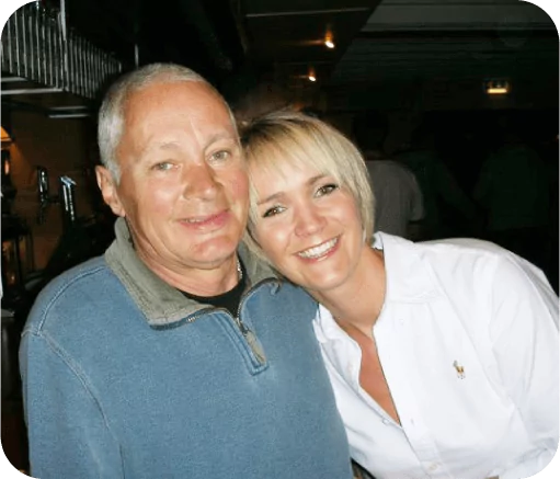 A smiling woman in a white shirt leans against a smiling older man in a blue sweater indoors.
