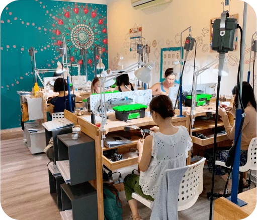 People working at jewelers benched in a colorful, decorated room with art on the walls and various jewelry making tools on the tables.