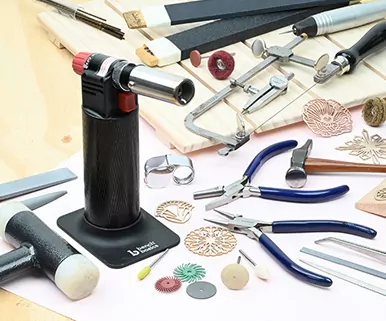 Jewelry-making tools on a table, including pliers, a torch, files, hammers, and various metal design pieces.