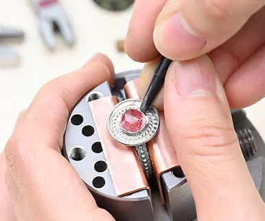 Close-up of hands setting a red gemstone into a metal jewelry piece using a small tool.