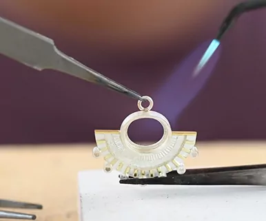 Close-up of a jeweler soldering a decorative silver jewelry piece with a blue flame.