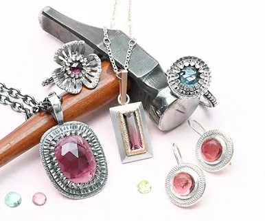 Silver jewelry with pink and blue gemstones displayed with a jewelers hammer and wood handle on a white background.
