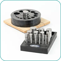 Metallic die punch set with circular base and various cylindrical punches on a block, displayed on a wooden platform.