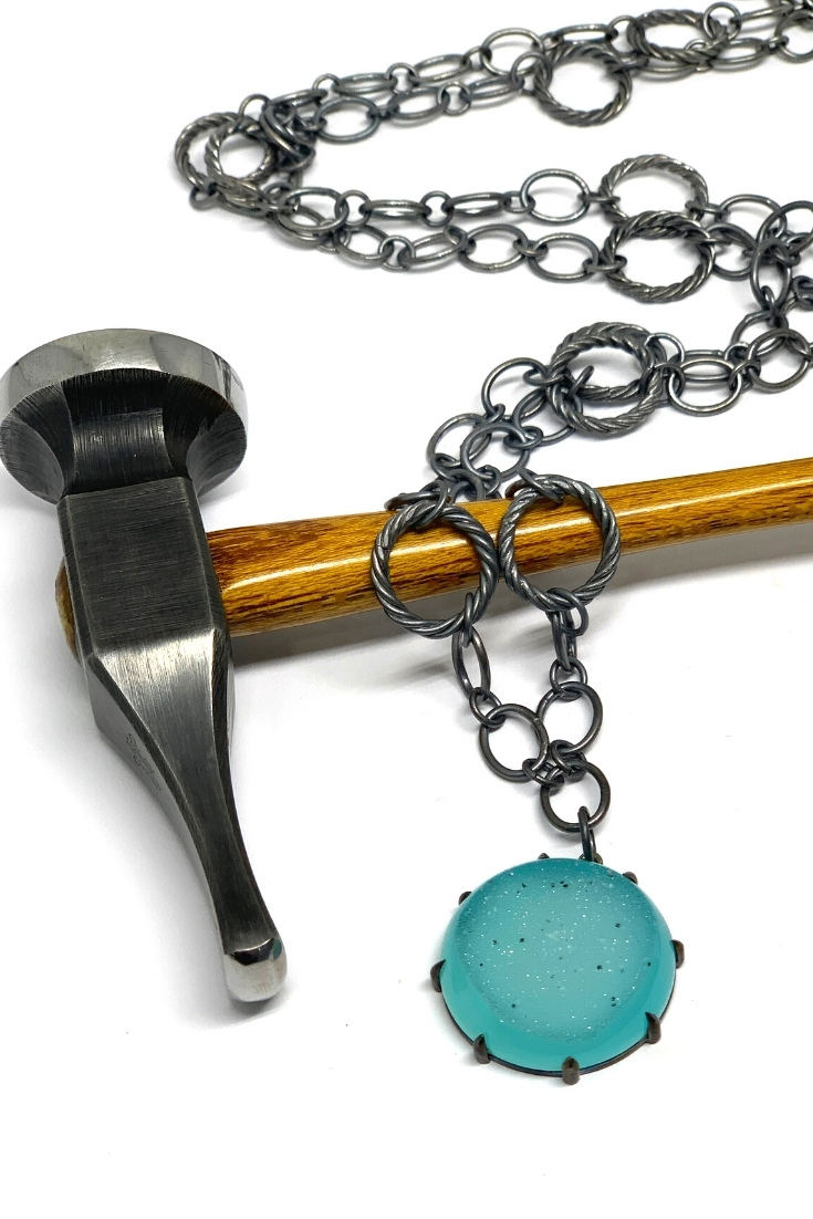Handmade sterling silver prong set pendant laying over a jewelers chasing hammer