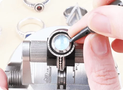 Person working on a gemstone ring held in a vise, with other rings nearby.