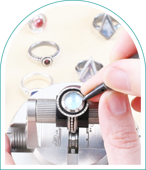 Hands crafting a ring with a blue gemstone in a vise, surrounded by various finished rings on a table.