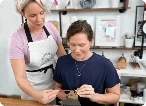 Two women crafting jewelry; one seated working, the other standing and guiding. Shelves with supplies in the background. From a class on how to make a ring with beaded wire.
