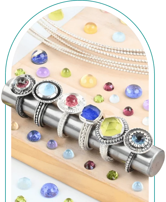 A row of colorful gemstone rings displayed on a metal rod with scattered loose stones in the background.