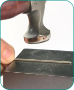 Hand holding a metal wire on an anvil, with a hammer poised above.