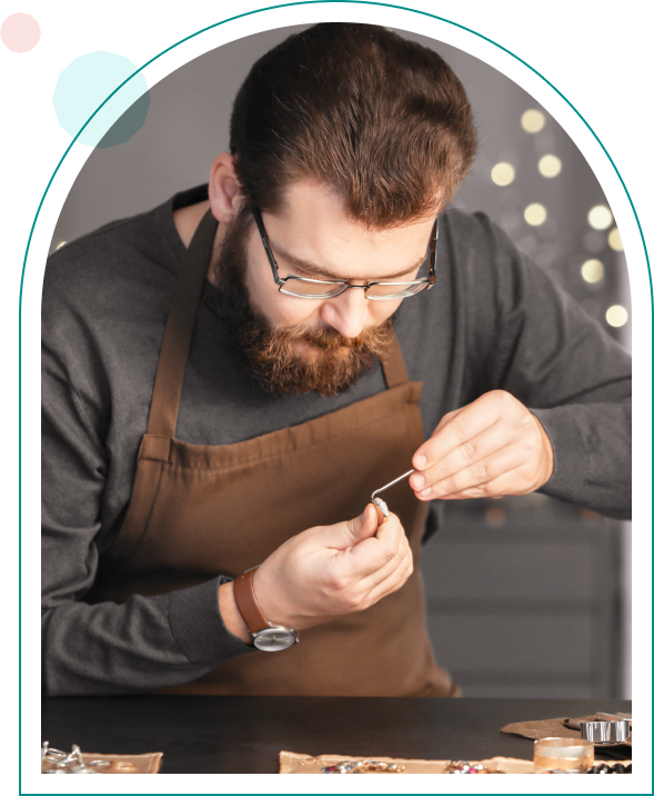 Bearded man in glasses and apron intricately crafting jewelry with pliers at a workbench.