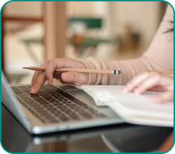 Person typing on a laptop with one hand, holding a pencil over a notebook with the other hand.