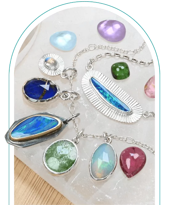 Colorful gemstone necklaces and pendants laid out on a light surface with various loose gemstones around them.  From a class on bezel setting for beginners.