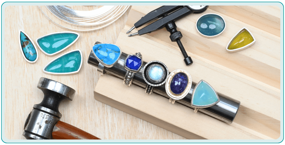 Jewelry-making tools and colorful gemstone rings on a wooden surface.From a class on how to bezel set a gemstone.