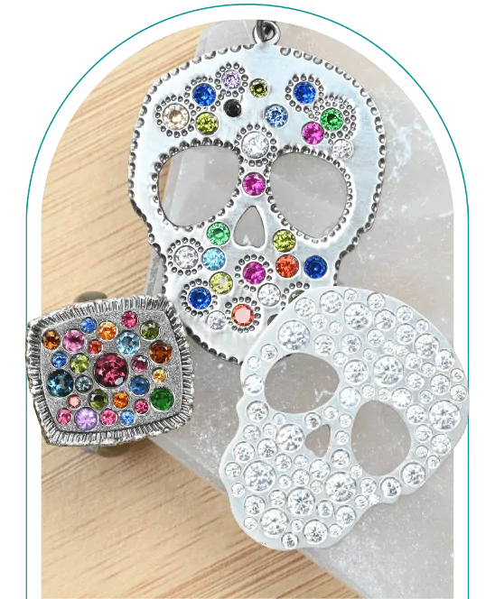 Decorative skull pendants and rings with colorful flush set gems, displayed on a wooden and marble surface. From a class on how to flush set stones.