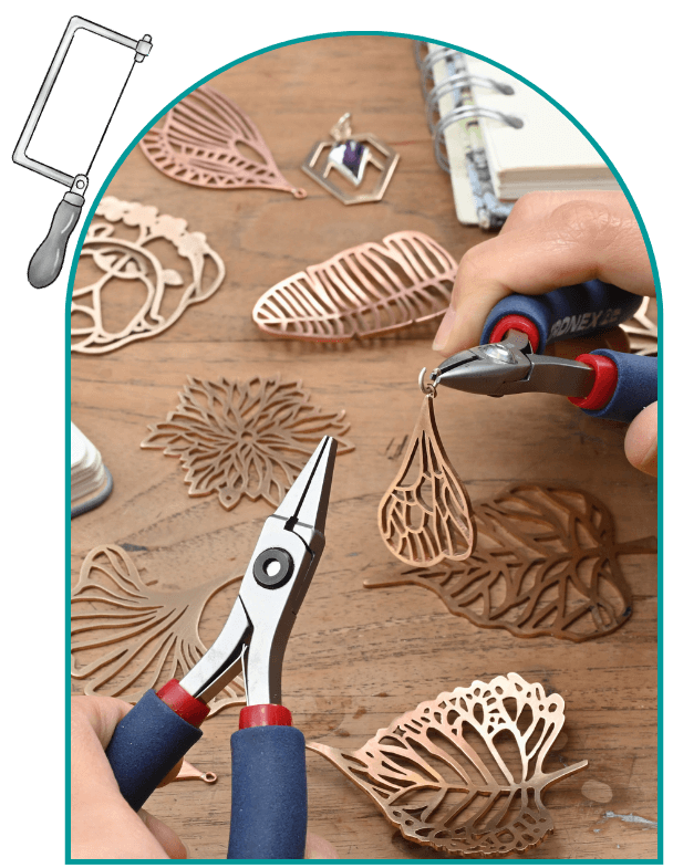 Two hands use jewelry making pliers on intricate metal leaf designs on a wooden table. From a class on how to use a jewelers saw.