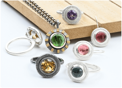 A collection of handmade gemstone rings and a chain necklace displayed on a wooden surface. From a class on making a halo ring.