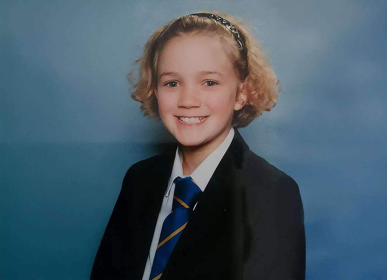 Photo of Lucy walker at age 11 with blonde curls and a dark headband in her school uniform.